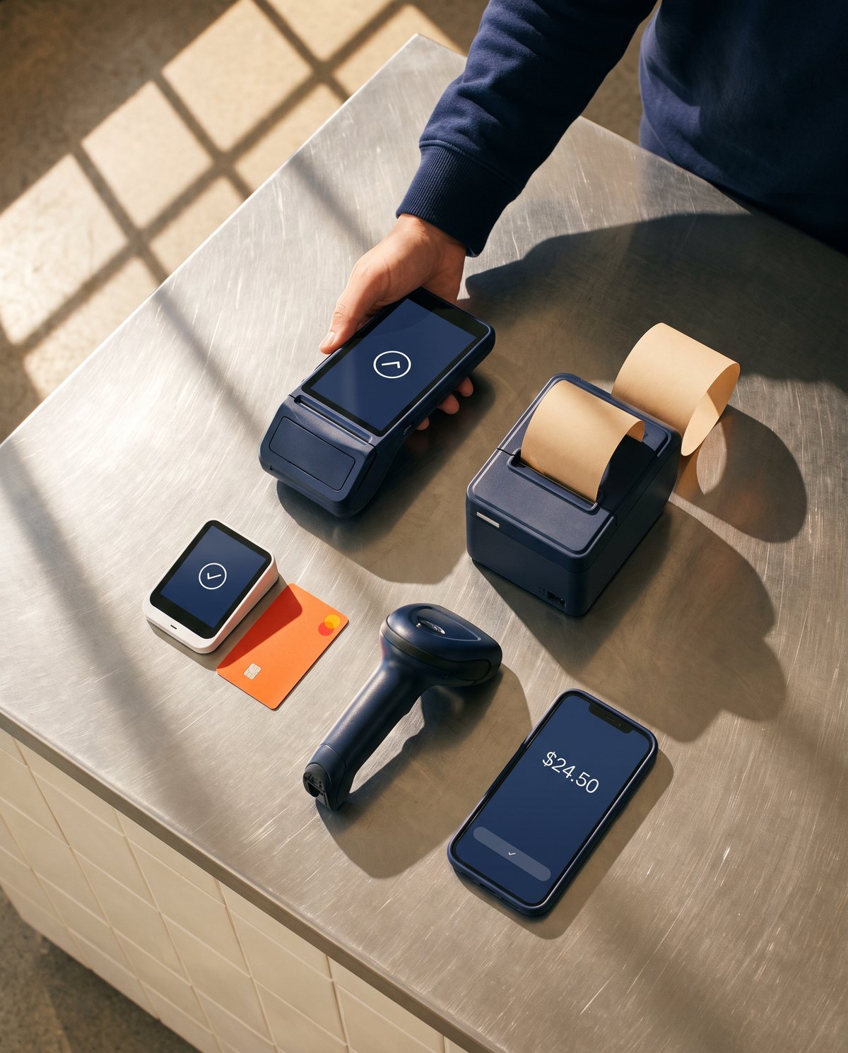 POS equipment laid out on a steel counter: a handheld card terminal, a small card reader, a receipt printer, a barcode scanner and a phone.